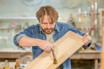 Man applies varnish on wooden board with paintbrush in  carpentry workshop