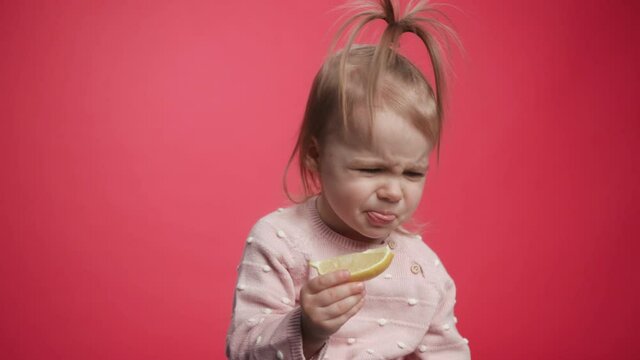 One Cute Beautiful Baby Eating a Lemon. A Child Tastes the Raw Lemon. Tiny Playful Girl Eats Fruits. Young Kid Holding Eat Tasty. Fun Expression Emotion Faces Kids from Eating Sour Lemon. Bright Look