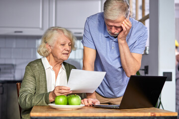 senior couple checking bills while managing accounts on home banking app. casual gray-haired man and woman using laptop while looking at invoice and plan the budget to save