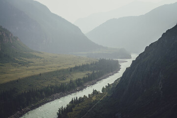 Misty mountain landscape with wide mountain river. Dark green gloomy scenery with confluence of two...