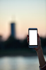 Young woman holding cellphone with blank white screen in urban city surroundings.