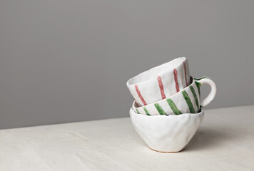 White ceramic cups with colorful stripes stack on table with linen tablecloth and gray wall...