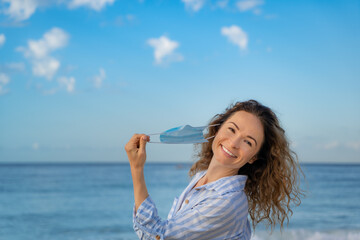 Fototapeta na wymiar Happy woman wearing medical mask outdoor against blue sea and sky background