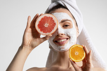 Smiling pretty young woman in towel with natural mask on face holding orange and grapefruit...