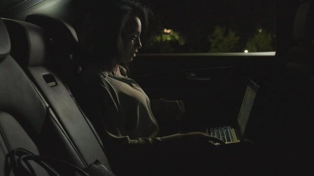 Medium Side-view Footage Of Mixed Race Business Lady Having Video Conference On Laptop Sitting At Backseat Of Modern Car With Black Leather Interior At Night, Talking To Colleagues