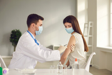 Obraz premium Male nurse or doctor in medical face mask giving injection to female patient during vaccination campaign at health center. Young woman getting flu or Covid-19 vaccine shot at the clinic or hospital