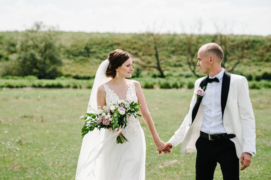 Bride and groom walking and holding bouquet of flowers and greens, greenery on nature. Romantic couple newlyweds outdoors. Wedding ceremony.