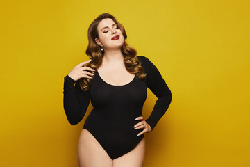 Plus-size model girl with bright makeup in black bodysuit posing at the yellow background. Young plus size woman in black bodysuits isolated on the yellow background