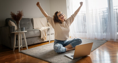 Pretty curly happy young woman sitting on floor at home in front of laptop with victory gesture...