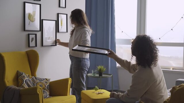 Guy with long curly hair helps young woman hang different frames with coloured dried leaves showing and sitting on sofa in new flat against window