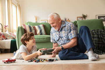 Elderly smiling grandfather giving little funny grandson present on holiday on warm floor among toys