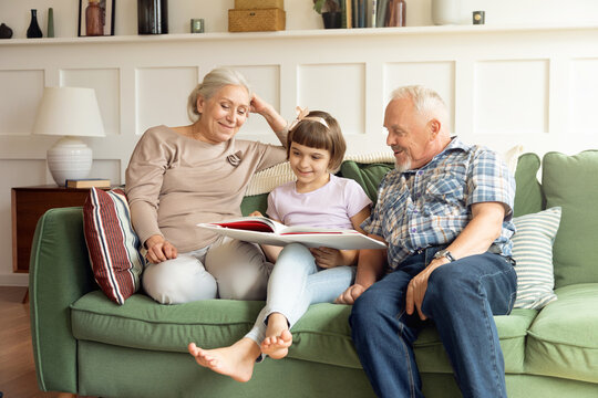 Happy 60s grandmother and grandfather sit resting on sofa at home read book with cute small granddaughter. Caring smiling grandparents help studying to little girl child. Education concept