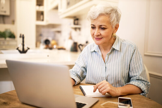 Creative mature female freelancer sitting in front of generic laptop making research, writing new post or article for her online blog on social media. Attractive retired woman using portable computer