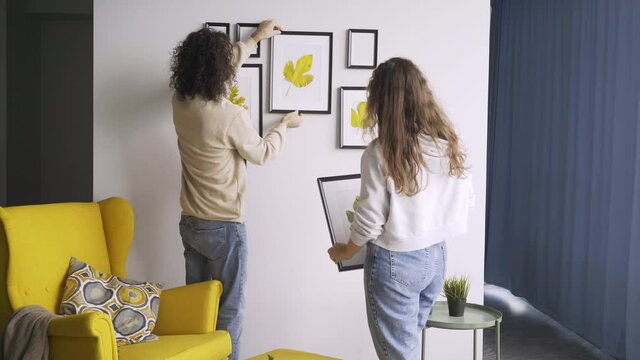 Romantic couple in jeans and pullovers hangs different sized wooden frames with coloured dried leaves on wall decorating new flat backside view