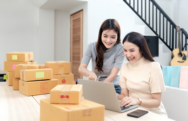 Two Asian women are looking at their laptop computers to check orders, with cardboard boxes and shopping bags all around, selling products online from home. being a new normal online business