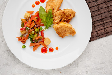 stewed vegetables with herbs and chicken steaks in batter on a white plate with drops of ketchup on a gray concrete background top view, food menu with a place to copy the text