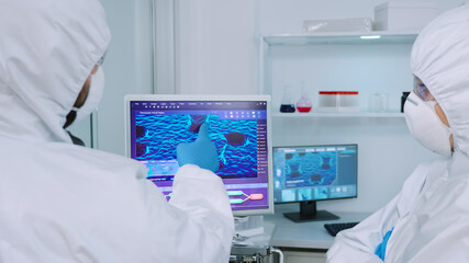 Scientists coworkers in coverall discussing about virus evolution in equipped laboratory pointing on computer desktop. Doctors examining vaccine evolution using high tech researching diagnosis