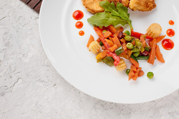 stewed vegetables with herbs and chicken steaks in batter on a white plate with drops of ketchup on a gray concrete background top view, food menu with a place to copy the text