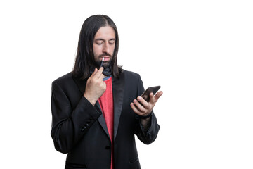 A man with long hair and a beard with an electronic cigarette with sticks and a phone in his hands on a white background. He is wearing a black jacket and a red T-shirt and a bow tie around his neck.