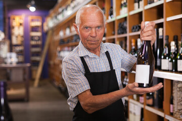 Portrait of confident elderly male owner of wine shop offering to buy wine bottle ..