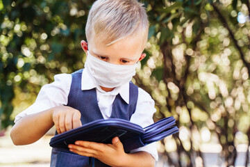 Young student of first grade stands in the schoolyard. Took out pencil from the pencil case. Wearing protective medical mask due to new safety requirements during the Covid-19 coronavirus pandemic