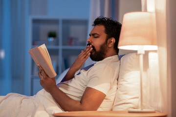 people, bedtime and rest concept - tired indian man reading book and yawning in bed at home at night - obrazy, fototapety, plakaty