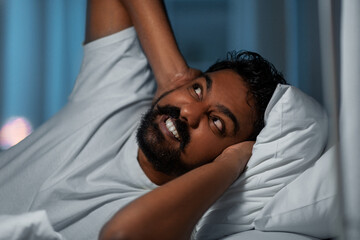 people, bedtime and rest concept - annoyed indian man lying in bed at home and covering ears at...