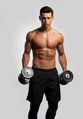 Front image of a confident young man shirtless torso portrait training with dumb-bell, isolated grey background. Vertical view.