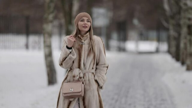 A beautiful blonde in a fur coat and a hat, she walks on the street, walks past the trees, poses. The model is located in the city park park in winter.