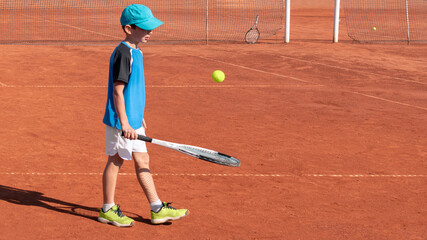 Child tennis player plays with ball and racket on red clay court. Childrens sports and physical activity concept. Kids fun sport game. Background, copy space