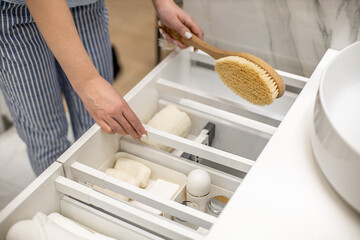 Happy young woman is holding wooden brush and putting into drawer together with bathroom amenities.