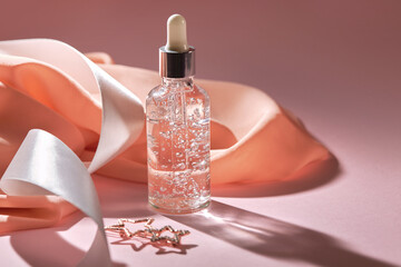 Anti aging serum in a glass bottle on a pink background with silk scarf, white satin ribbon nd star...