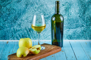 A glass of white wine and bottle with apple slices on blue background