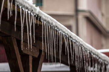 Icicles forming ice stalactites falling fron the edge of the eaves of gutters and roofs of a residential building during a cold winter afternoon after a wave of snow made the temperatures falling