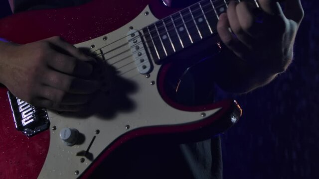 Rock musician plays a red white electric guitar in a dark studio against the background of falling rain drops. Close up of male hands play on strings with guitar pick. Slow motion.