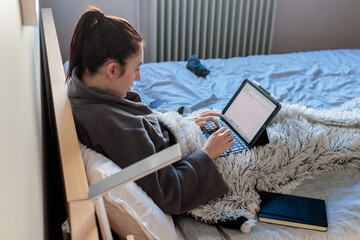 Mujer caucásica joven sentada en la cama de su habitación con ropa e pijama tapada con una manta usando una tablet estudiando en casa. 
