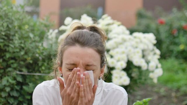 Girl sneezes into a napkin, because she is allergic to flowering during spring time on white chrysanthemums on background, hypersensitivity concept