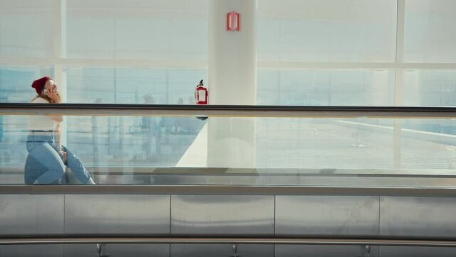 Clean simple shot of young female traveller sit on top of suitcase on moving walkway or travelator in empty airport terminal. Use smartphone, bored waiting for delayed or cancelled flight information
