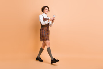 Fototapeta na wymiar Full length body size photo of cheerful schoolgirl using smartphone blogging smiling isolated on pastel beige color background