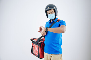 Young serious courier in helmet and medical mask carrying cooler bag and checking time on smartwatch