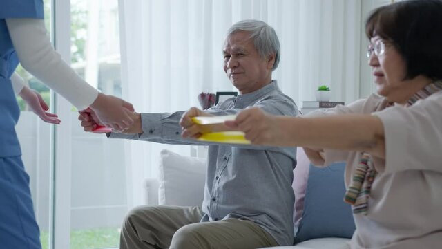 Young older senior asia citizen couple sit on sofa couch with in-home nursing care, assisted living, scrubs nurse use resistance band exercise for senior patient in physiotherapy treatment at home.