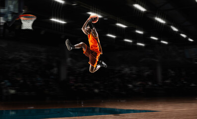 Fototapeta na wymiar In jump flight African-american young basketball player in action and motion in flashlights over dark gym background. Concept of sport, movement, energy and dynamic, healthy lifestyle. Arena's drawned