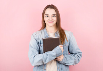 Portrait of beautiful young girl holding book and looking at camera isolated on pink wall background