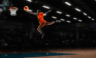 Leader. African-american young basketball player in action and motion in flashlights over dark gym background. Concept of sport, movement, energy and dynamic, healthy lifestyle. Arena's drawned.