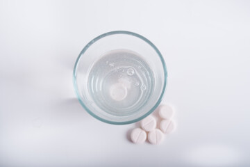 A glass of pure water on a white background, in which is a dissolving vitamin that forms bubbles. Next to it lie round pills. Disease treatment.