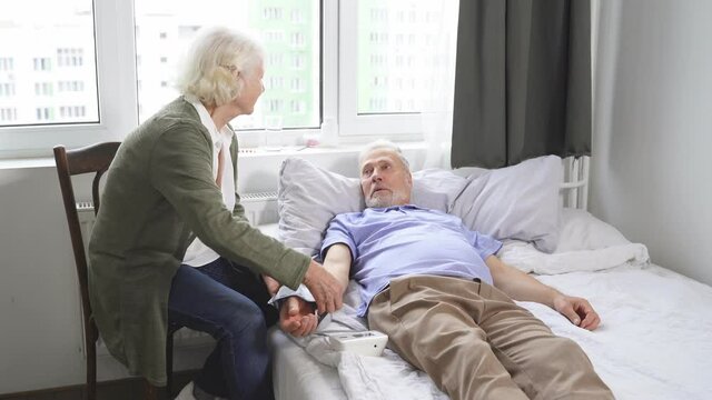 an elderly woman checks her husband's blood pressure with a blood pressure monitor. A man is lying at home in bed. A woman's concern for a man.