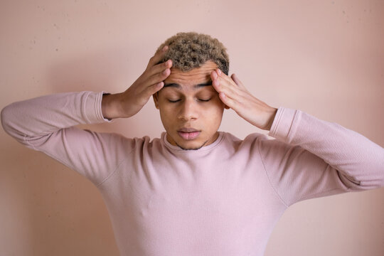 Young tired African American man holding head suffering from headache on early morning against pink kinfolk wall background