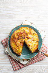 Homemade Spanish tortilla with one slice cut - omelette with potatoes on plate on white wooden rustic background top view. Traditional dish of Spain Tortilla de patatas for lunch or snack, overhead
