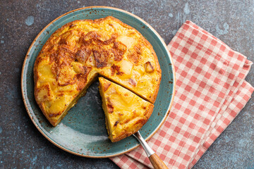 Homemade Spanish tortilla with one slice cut - omelette with potatoes on plate on stone rustic background top view. Traditional dish of Spain Tortilla de patatas for lunch or snack, overhead
