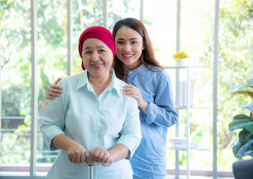 Asian Elderly Mother Wearing Red Headscarf Recover From Cancer Standing, Holding Walking Stick And Having Her Daughter By Her Side Embrace, Smile Very Happily. Cancer Or Leukemia Survivor Concept.